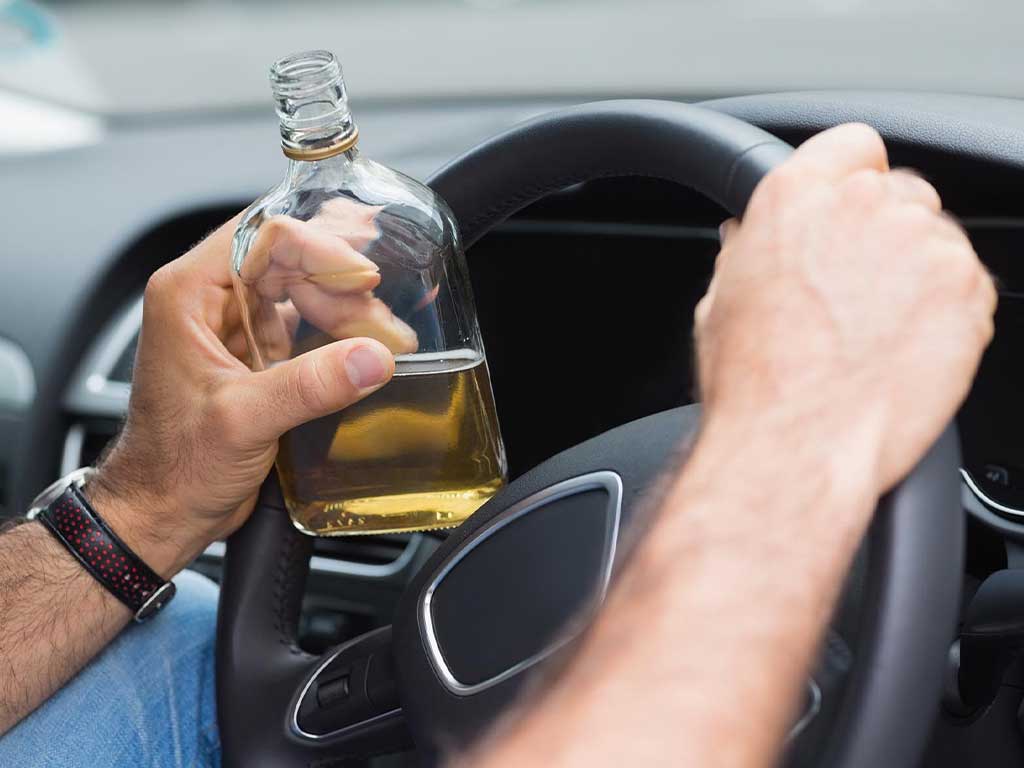 A man holding the steering wheel of a car while holding a bottle of alcohol