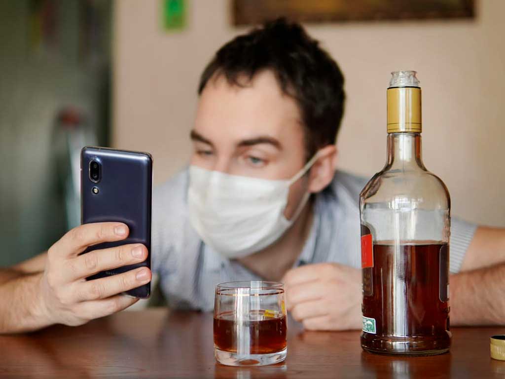 A man looking at his phone with a glass and bottle of alcohol on the table