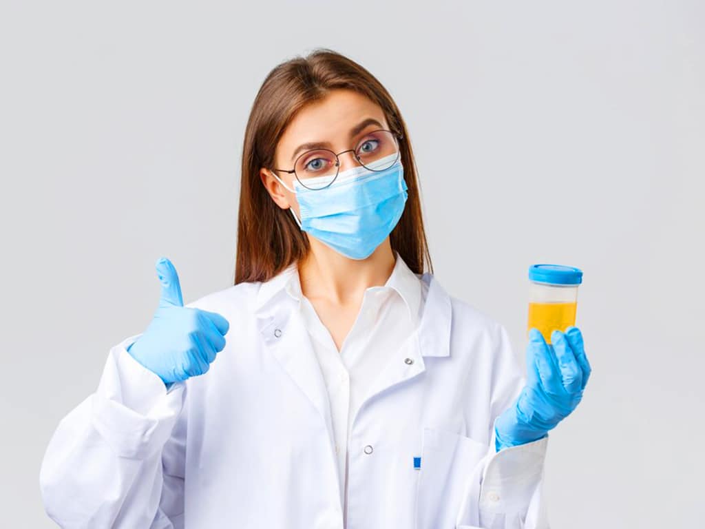 A female laboratory technician holding a urine sample cup and making the "okay" hand gesture