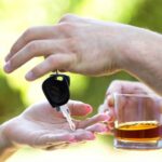A person holding a glass of alcohol on one hand and giving a car keys to another on the other hand